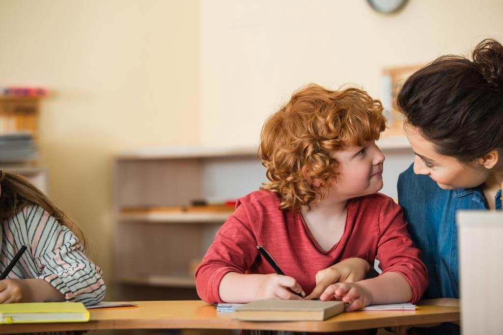 roodharige jongen kijkt naar leraar wijzend naar kopieerboek in de klas - Foto, afbeelding