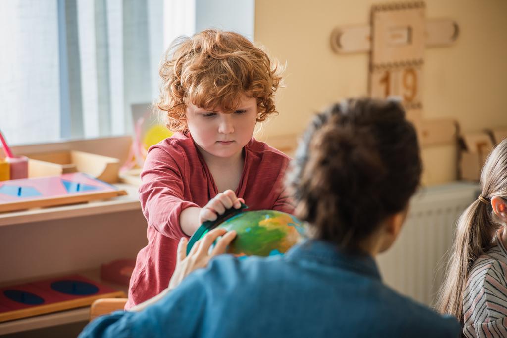 кудрявый мальчик смотрит на глобус рядом с размытыми учительницей и девочкой в школе Монтессори - Фото, изображение