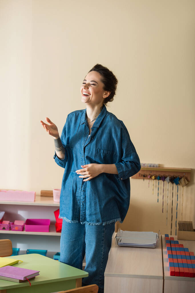 fröhliche Lehrerin im Jeanshemd lacht mit geschlossenen Augen in Montessori-Schule - Foto, Bild