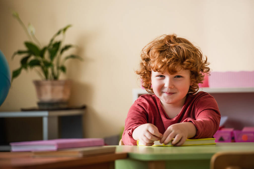 redhead and curly boy smiling at camera near blurred books in montessori school - Photo, Image