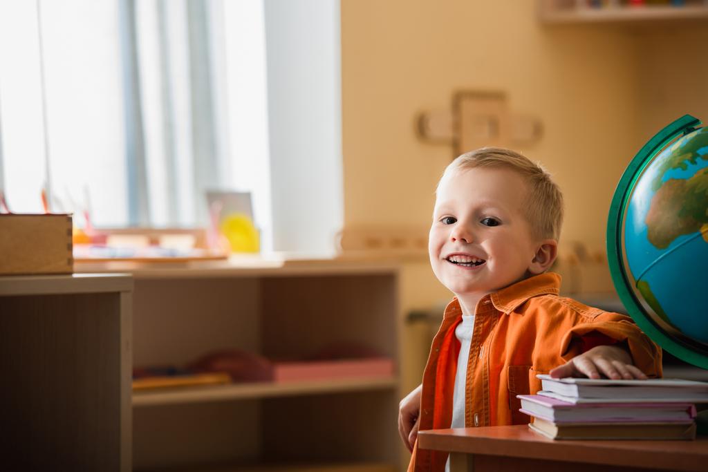 menino feliz sorrindo para a câmera perto do globo e livros na escola montessori - Foto, Imagem