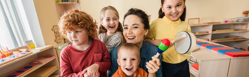 cheerful teacher looking at camera while playing music with interracial kids, banner - Photo, Image
