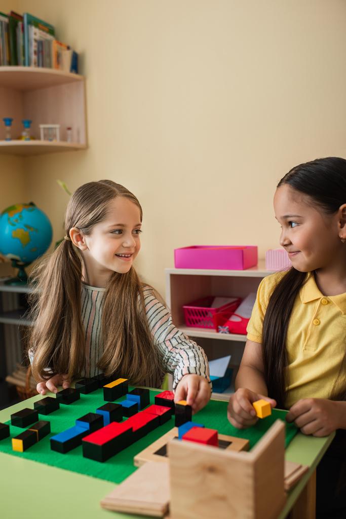happy interracial girls looking at each other while playing with wooden blocks in montessori school - Photo, Image