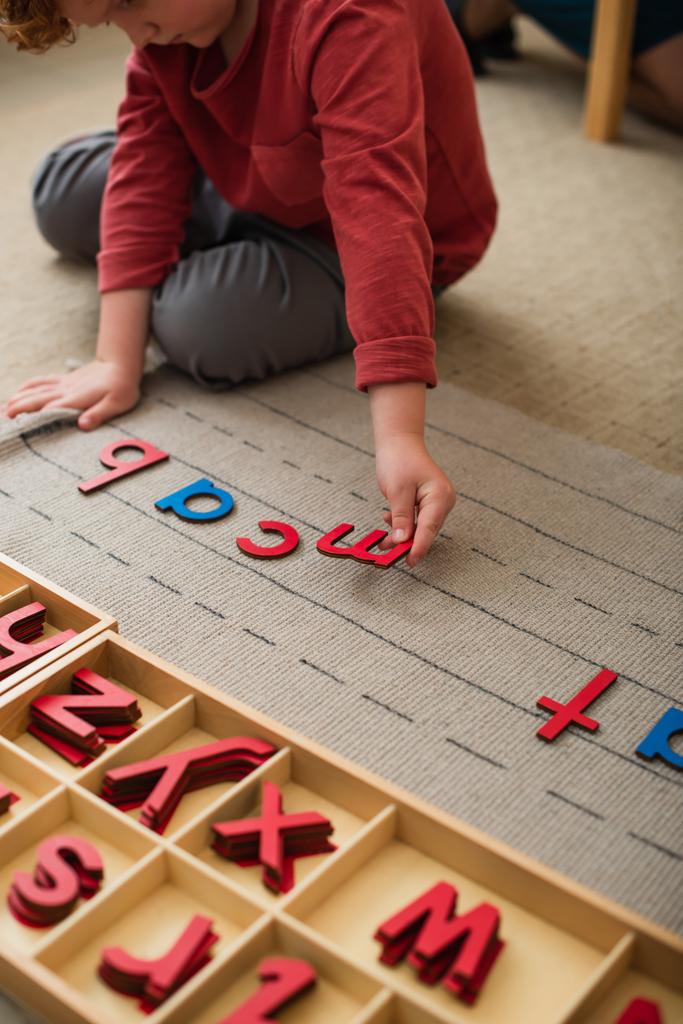 częściowy widok chłopca bawiącego się drewnianymi literami na podłodze w szkole montessori - Zdjęcie, obraz