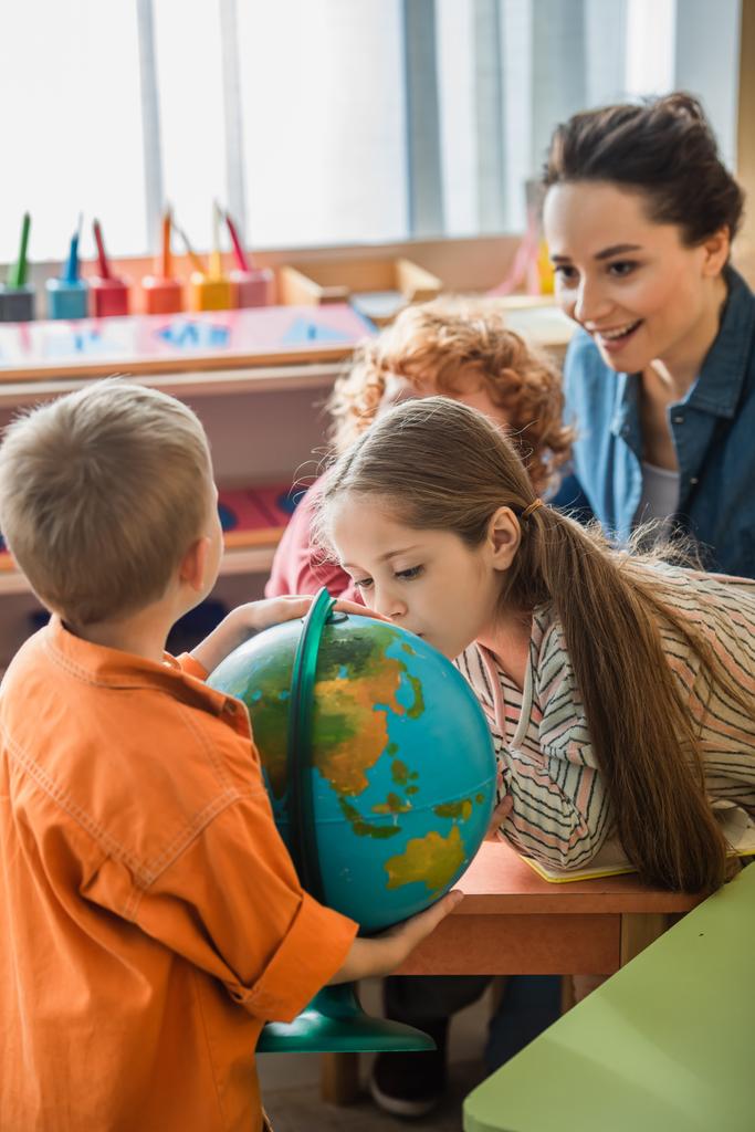 Любопытная девушка смотрит на глобус рядом с детьми и размытый учитель в классе - Фото, изображение