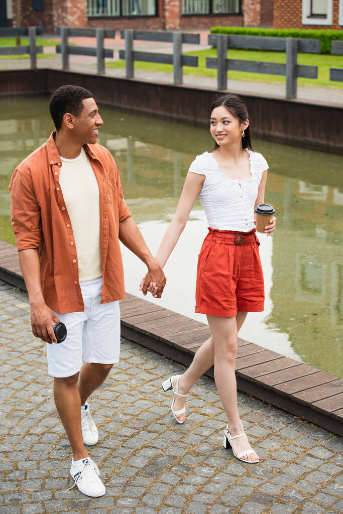 stylisches multiethnisches Paar mit Pappbechern Händchen haltend beim Spaziergang am Wasser in der Stadt - Foto, Bild