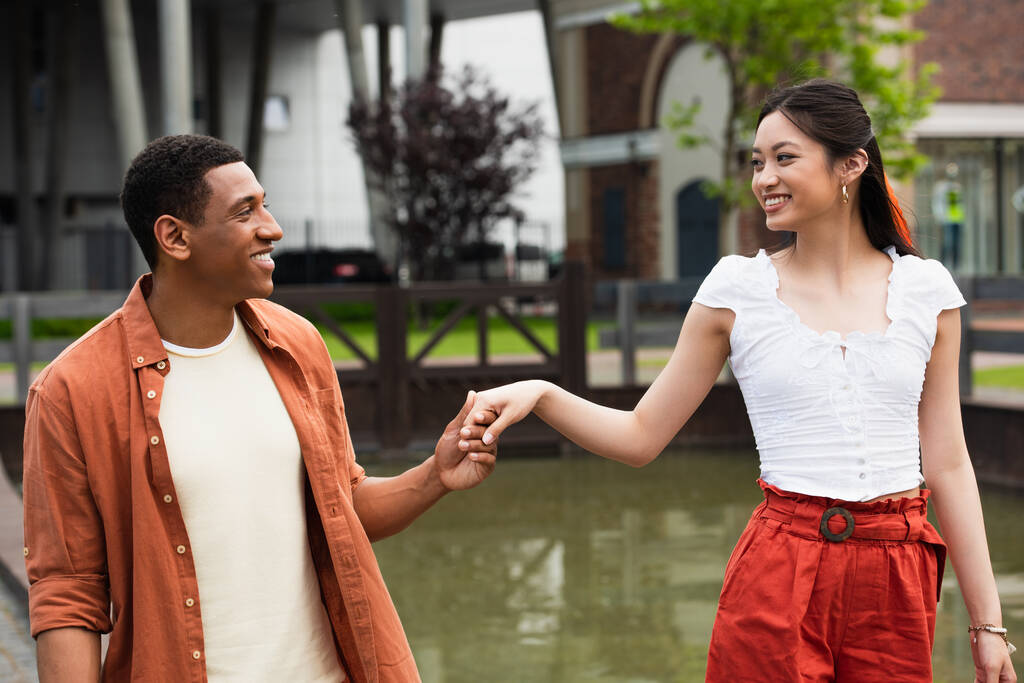 joyful multiethnic couple looking at each other and holding hands while walking near water - Photo, Image