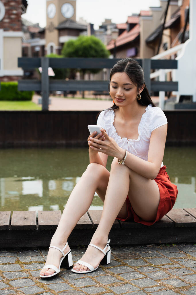 улыбающаяся азиатка, сидящая на границе возле воды и использующая смартфон - Фото, изображение