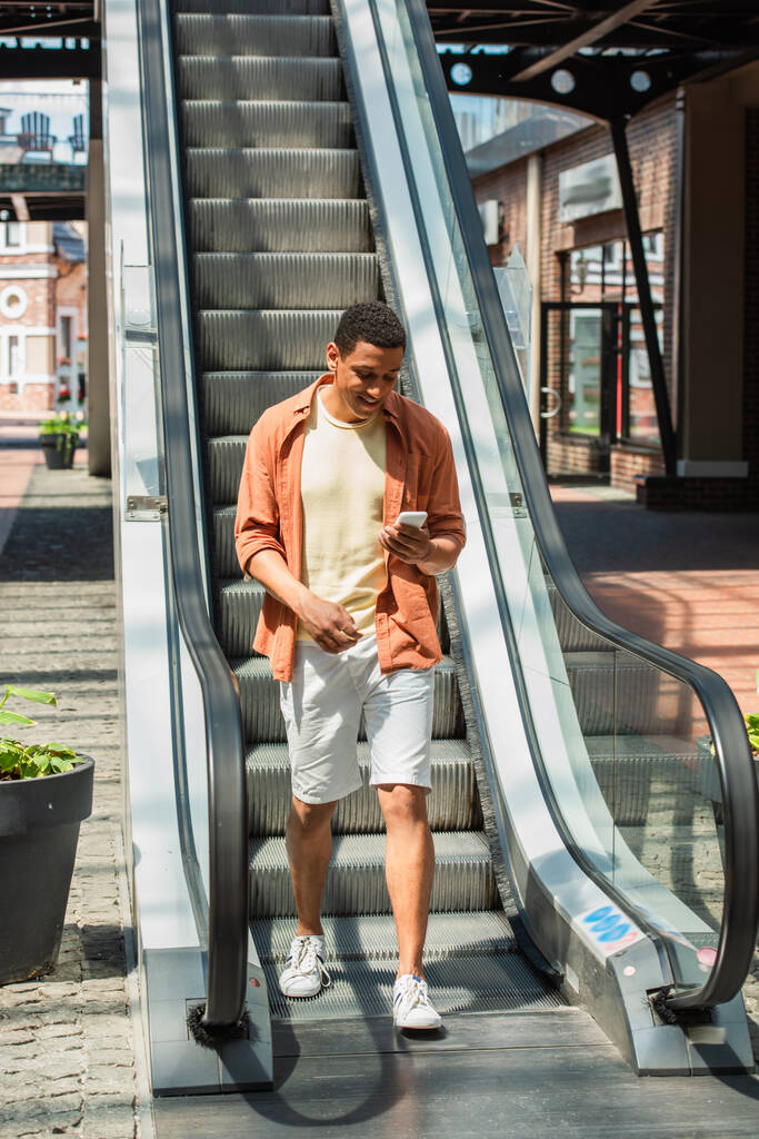 allegro uomo africano americano in pantaloncini utilizzando il telefono cellulare vicino alla scala mobile - Foto, immagini