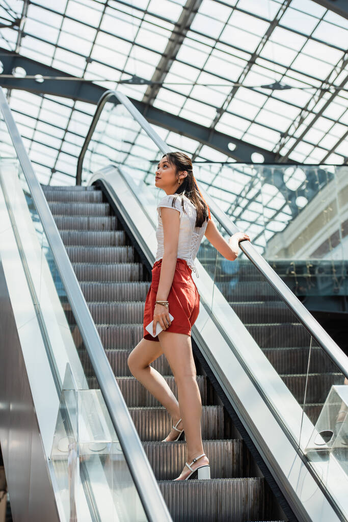 junge Asiatin hält Smartphone in der Hand, während sie auf Rolltreppe wegschaut - Foto, Bild
