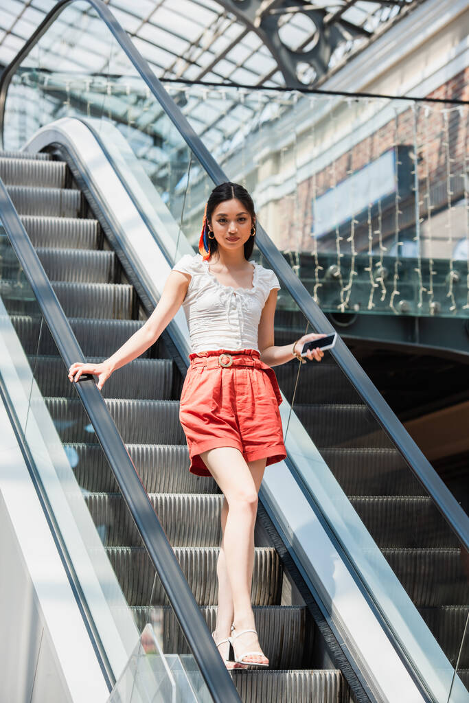 junge Asiatin in kurzen Hosen blickt mit Smartphone auf Rolltreppe in die Kamera - Foto, Bild