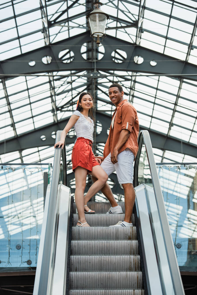 Yürüyen merdivende kameraya gülümseyen şık yaz kıyafetleri içindeki ırklar arası çiftin düşük açılı görüntüsü - Fotoğraf, Görsel