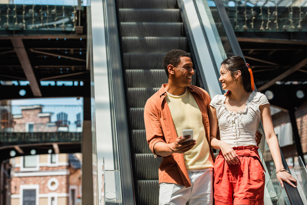 stylish african american man with smartphone smiling at asian girlfriend near escalator - Photo, Image