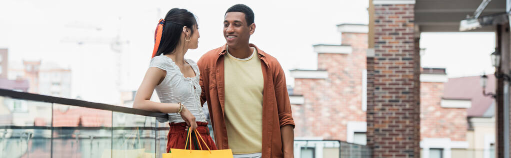 Afro-Amerikalı mutlu bir adam alışveriş çantası ve pankartıyla güzel bir Asyalı kadına bakıyor. - Fotoğraf, Görsel