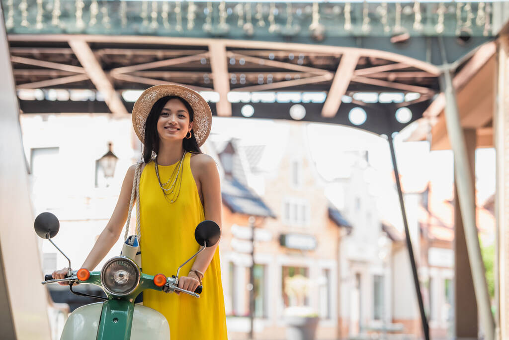feliz asiático mujer en amarillo sundress y paja sombrero sonriendo cerca scooter - Foto, Imagen
