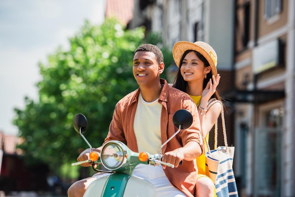 alegre asiático mujer tocando paja sombrero mientras cabalgando scooter con africano americano novio - Foto, Imagen