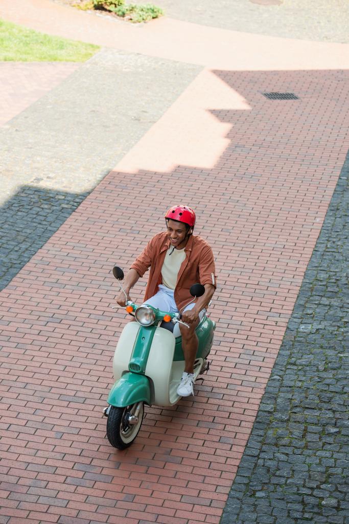 vue grand angle de l'homme afro-américain en hardhat équitation scooter sur la chaussée - Photo, image