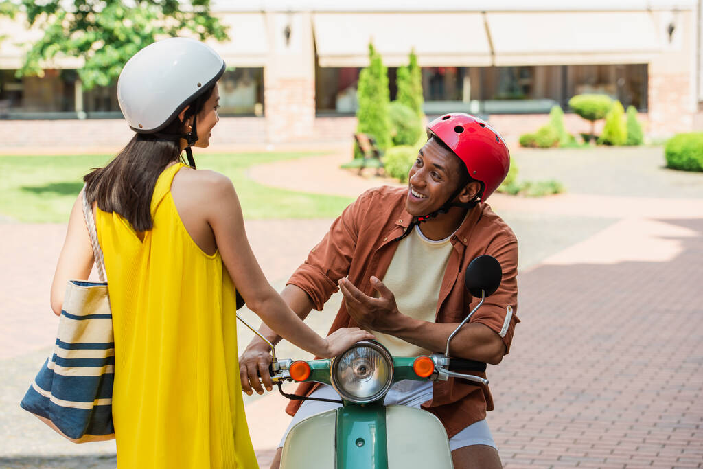 улыбающийся африканский американец на скутере разговаривает со стильной азиаткой на улице - Фото, изображение