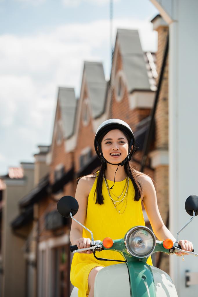 jeune femme asiatique en hardhat souriant tout en chevauchant scooter en ville - Photo, image