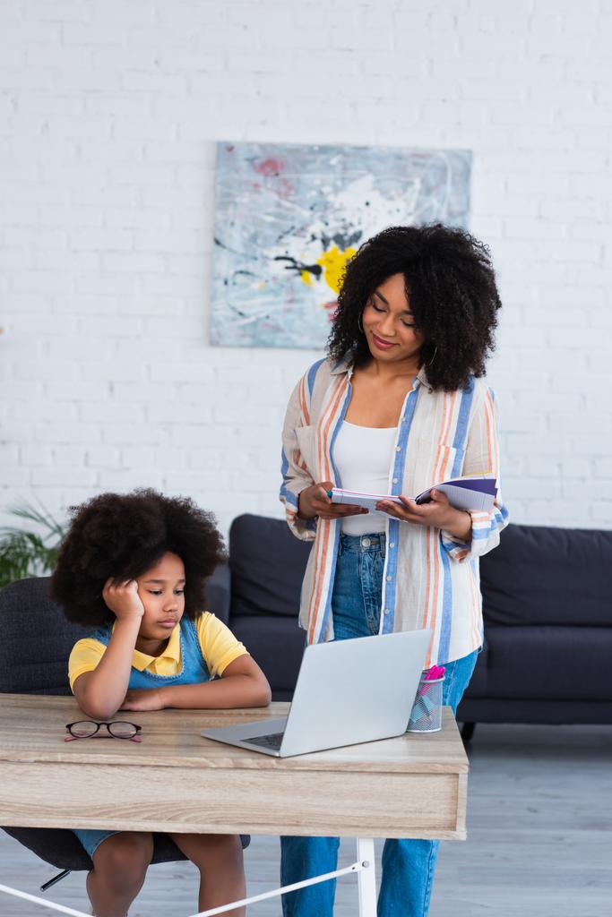 Mosolygó anya notebook nézi afro-amerikai gyerek használ laptop otthon  - Fotó, kép