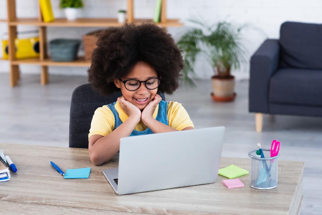 Afrikanisches Kind mit Brille blickt auf Laptop neben Schreibwaren auf Tisch  - Foto, Bild