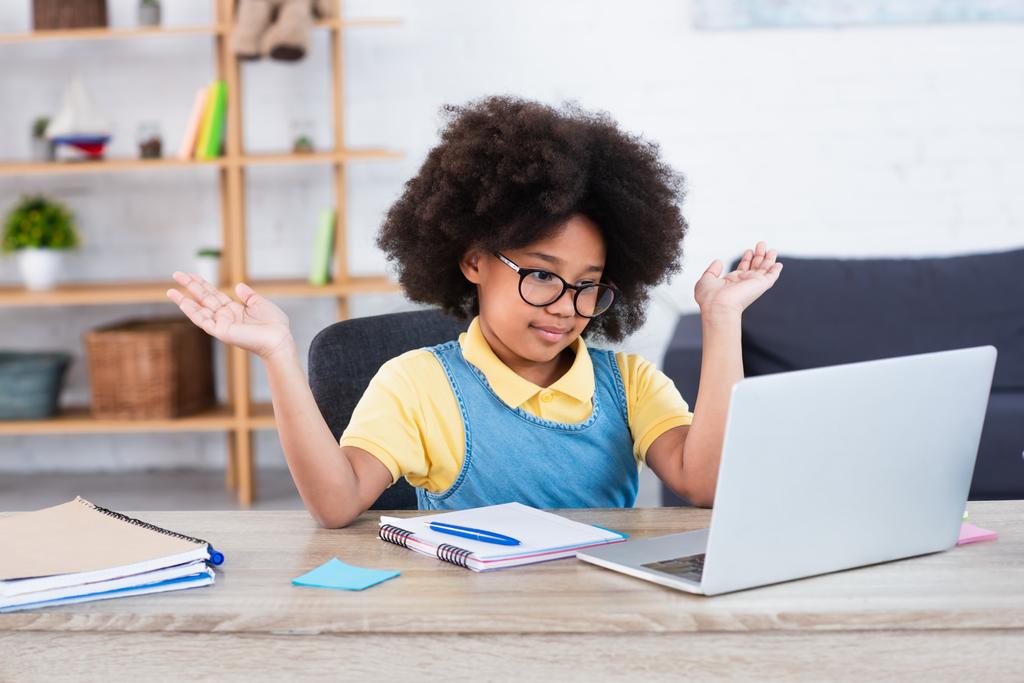 African american girl in eyeglasses pointing with hands at laptop near notebooks at home  - Photo, Image