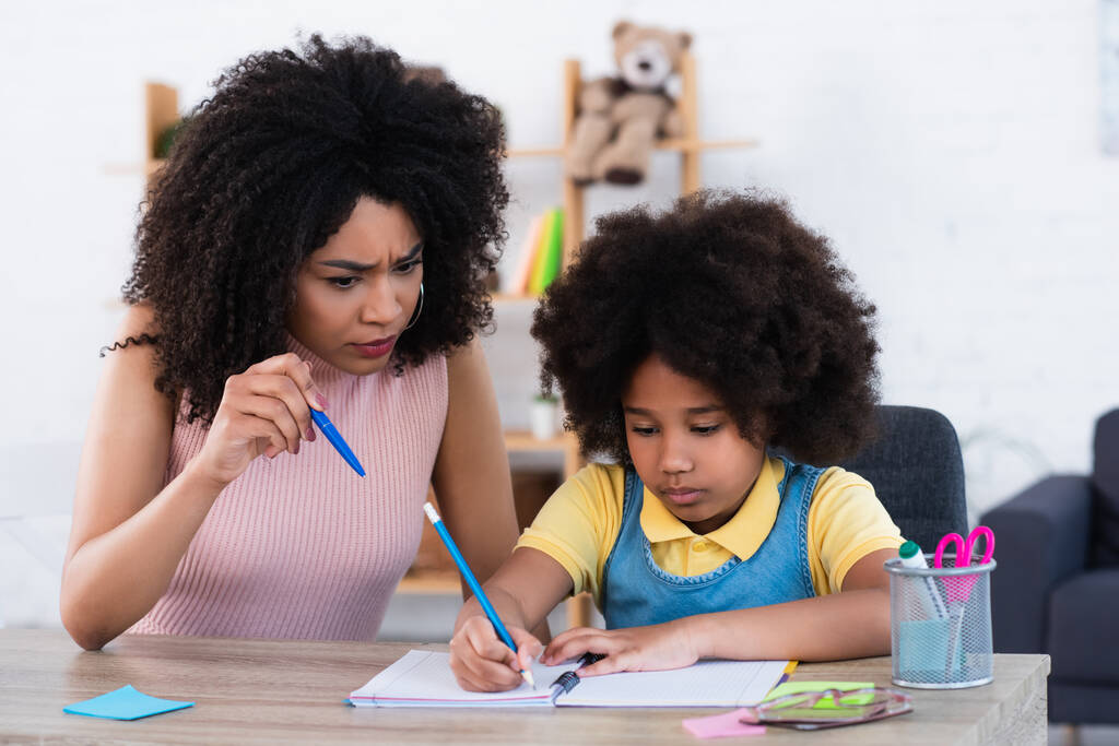 Mère afro-américaine en colère pointant avec un stylo près de sa fille écrivant sur un carnet à la maison  - Photo, image