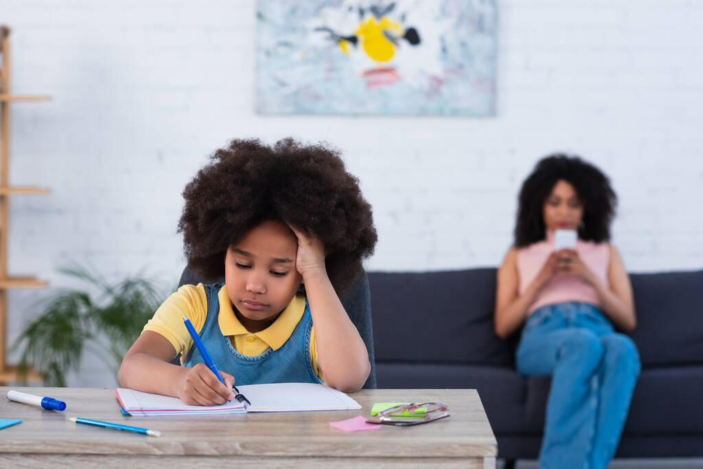 Afrykańskie amerykańskie dziecko piszące na notatniku w pobliżu rozmytej matki ze smartfonem w domu  - Zdjęcie, obraz