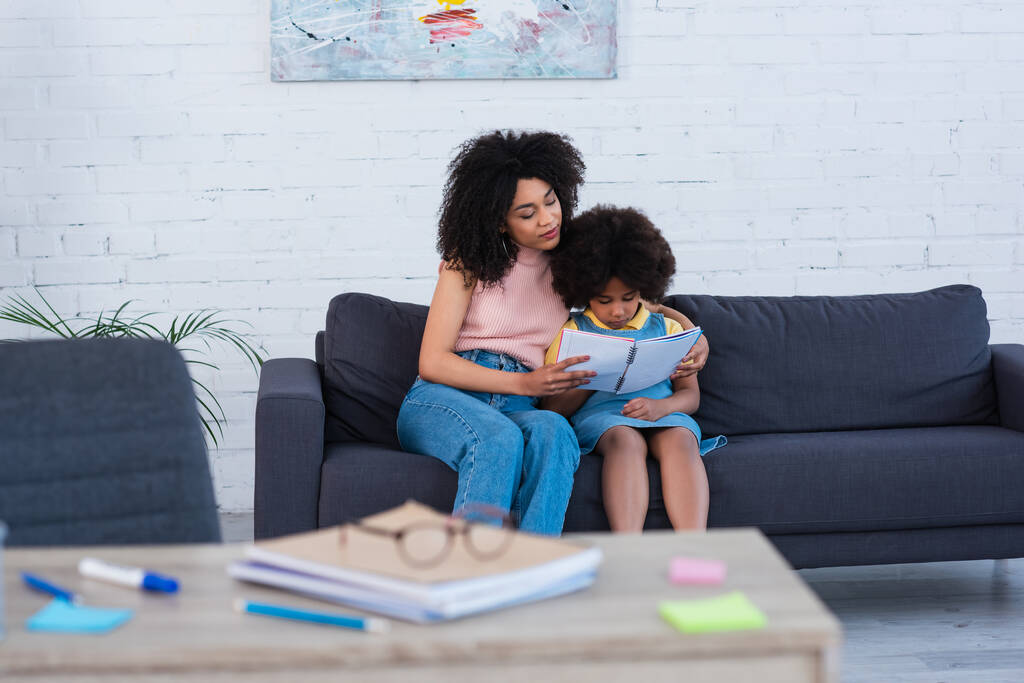 Африканская американская мать держит блокнот рядом с дочерью на диване  - Фото, изображение