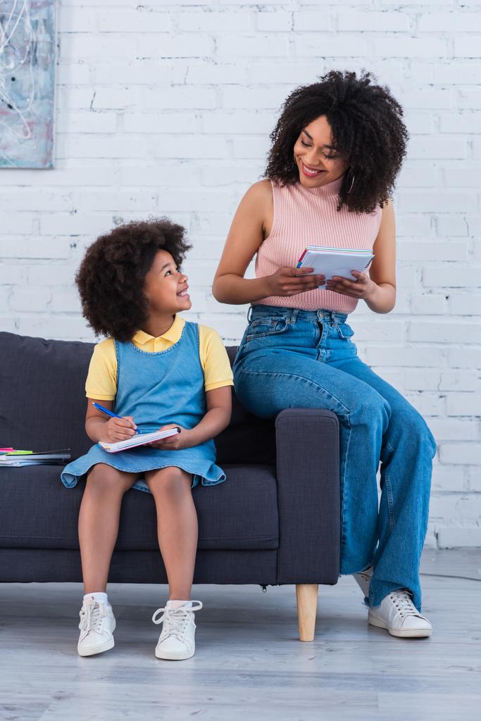 Souriant enfant afro-américain qui regarde sa mère avec un cahier à la maison  - Photo, image