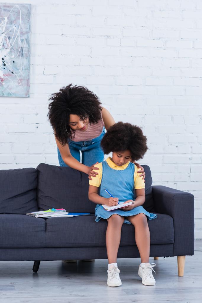 Femme afro-américaine embrassant enfant écriture sur ordinateur portable sur canapé  - Photo, image