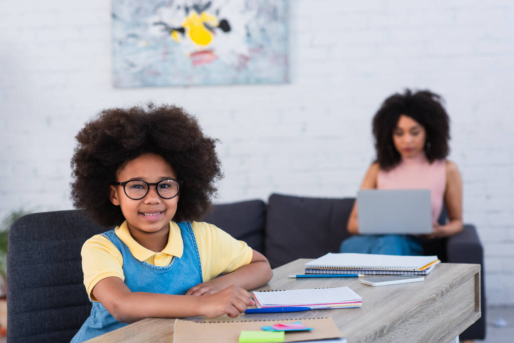 Mosolygó afro-amerikai gyerek szemüvegben ül közel kellékek és elmosódott anya laptoppal  - Fotó, kép