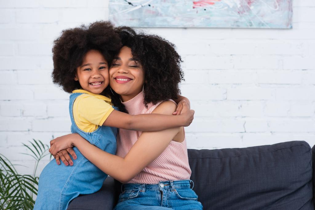 Mãe com os olhos fechados abraçando a criança americana africana positiva na sala de estar  - Foto, Imagem