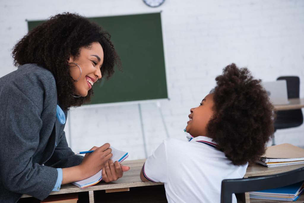 Professora afro-americana sorridente escrevendo em caderno perto de aluno em sala de aula  - Foto, Imagem