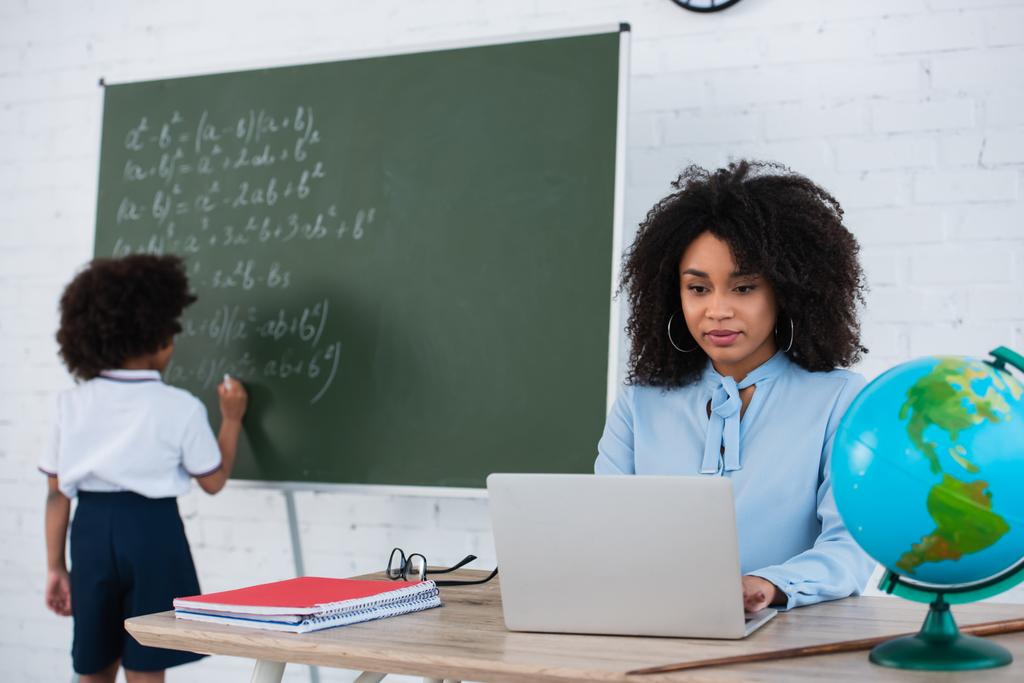 Professeur afro-américain utilisant un ordinateur portable pendant que l'élève écrit sur un tableau noir sur fond flou  - Photo, image