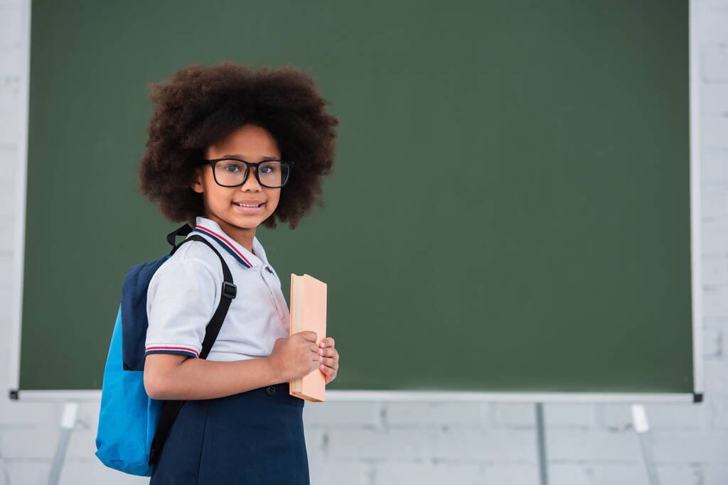 Niño afroamericano feliz con mochila y libro de pie cerca de pizarra en la escuela  - Foto, imagen