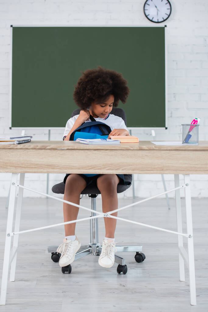 Afrikai amerikai iskolás lány hátizsákkal nézi könyv íróasztal osztályteremben  - Fotó, kép