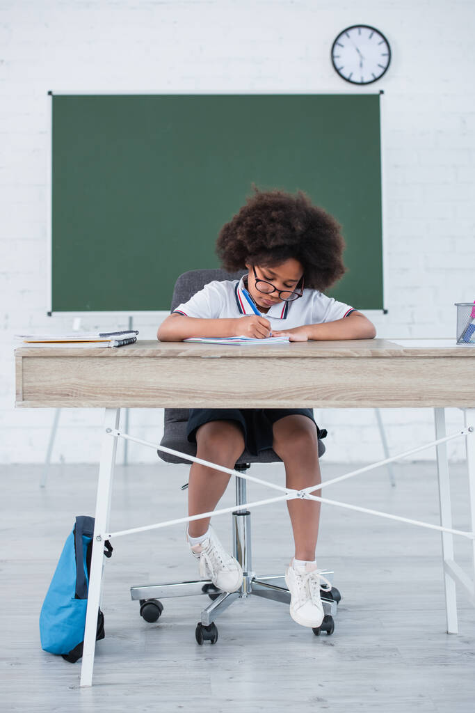 Afrikaans-Amerikaanse leerling in bril schrijven op notebook in de buurt van rugzak in de klas  - Foto, afbeelding