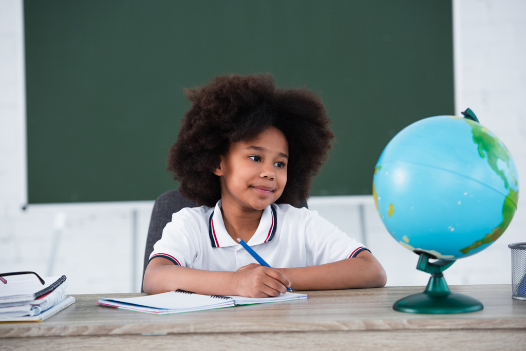glimlachende Afrikaans-Amerikaanse leerling schrijven op notebook in de buurt wazig globe op bureau  - Foto, afbeelding