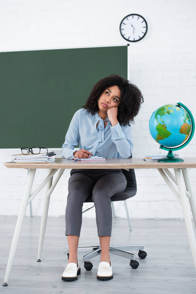 Pensive афроамериканський вчитель тримає ручку біля глобуса і канцелярські товари в класі
  - Фото, зображення