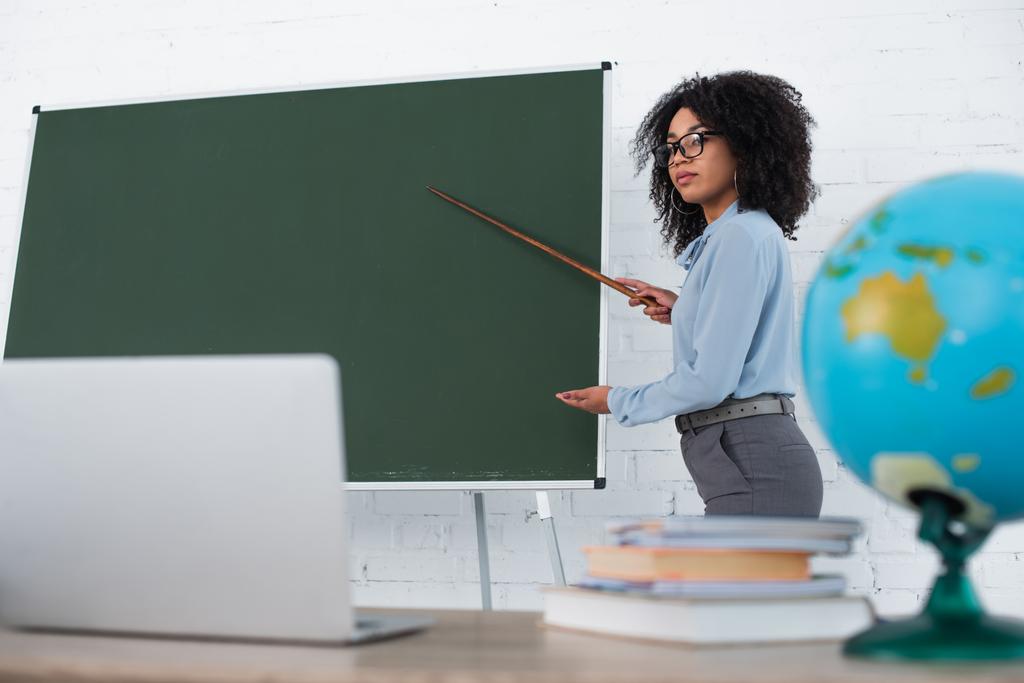 Afrykański amerykański nauczyciel wskazujący na tablicę w pobliżu zamazanego laptopa podczas lekcji online w szkole  - Zdjęcie, obraz