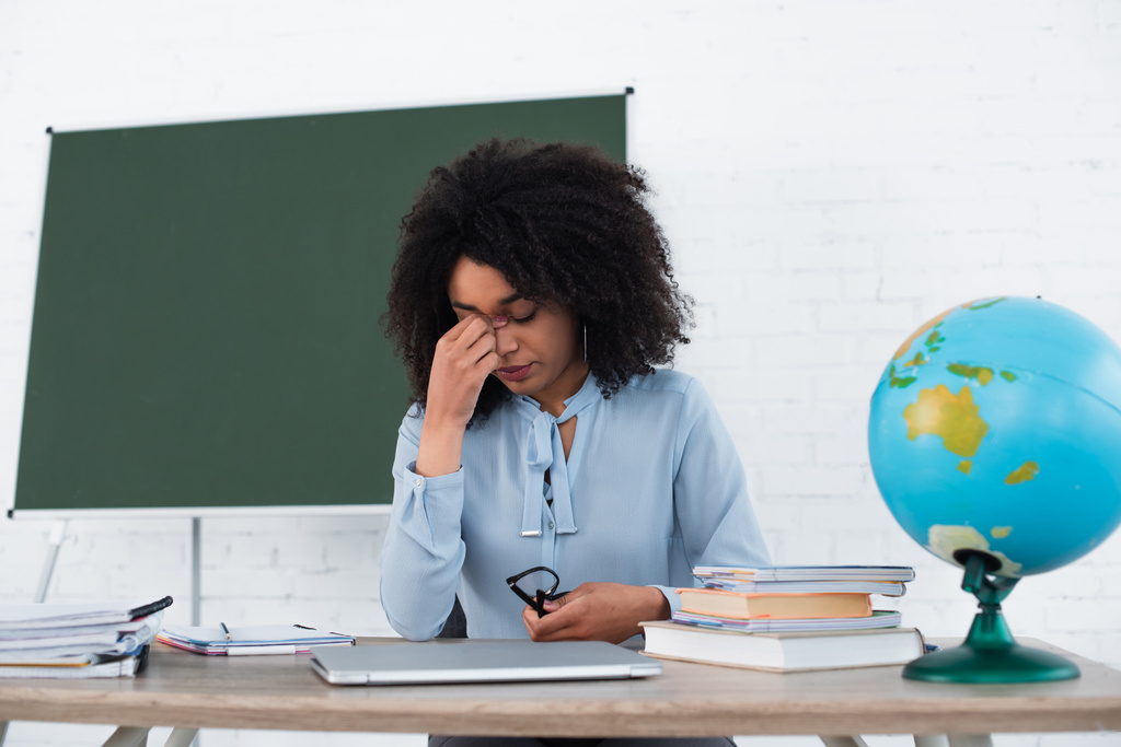 Втомлений афроамериканець тримає окуляри біля ноутбука і глобуса в класі.  - Фото, зображення