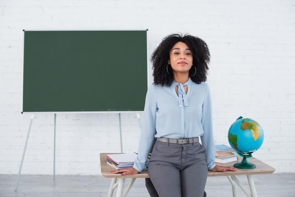 Junge afrikanisch-amerikanische Lehrerin steht neben Globus und Notizbüchern auf Arbeitstisch  - Foto, Bild
