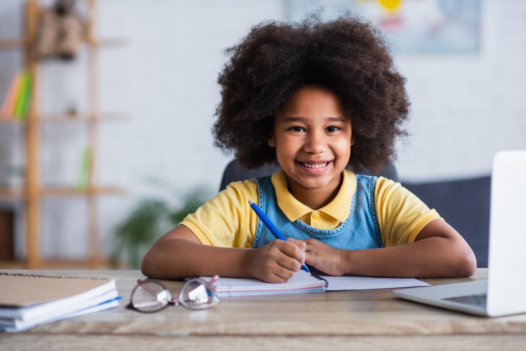Šťastná africká americká dívka psaní na notebook v blízkosti notebooku a brýle doma  - Fotografie, Obrázek