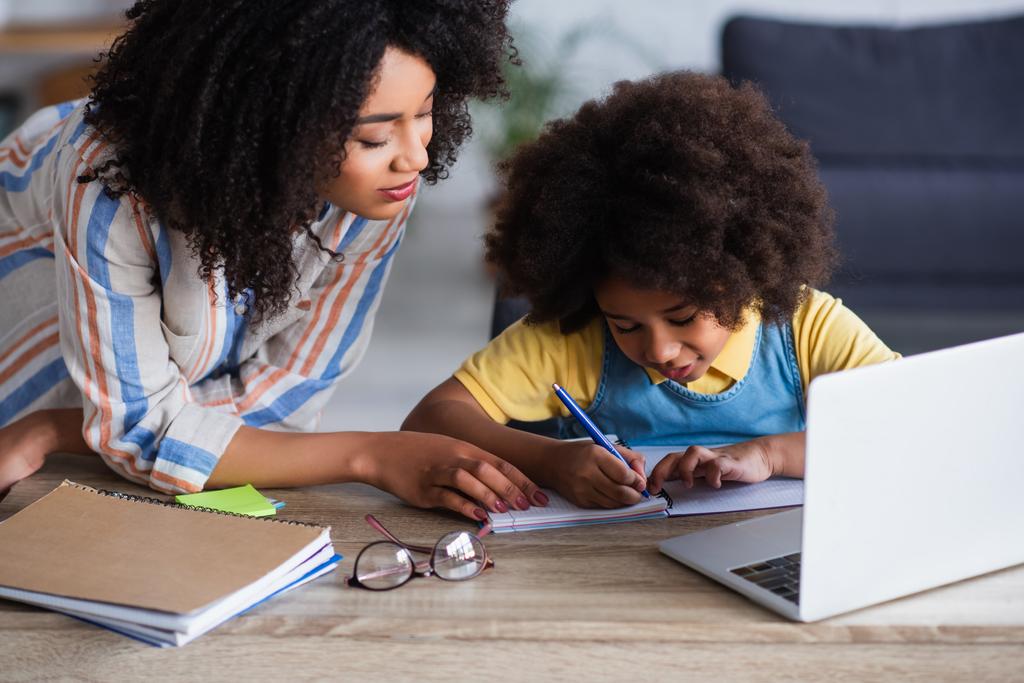Afroamerikanische Mutter steht neben Kind und schreibt während der Hausaufgabenbetreuung auf Notebook und Laptop  - Foto, Bild