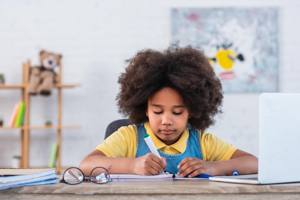 Африканська американська дитина з маркером на зошиті біля окулярів і розмитим ноутбуком.  - Фото, зображення