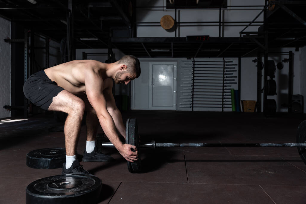 Молодой активный потный сильный мускулистый мужчина с большими мускулами кладет тяжелую железную пластину на штангу для тяжелой атлетики или тренировки по тяжелой атлетике, тренируя реальных людей - Фото, изображение