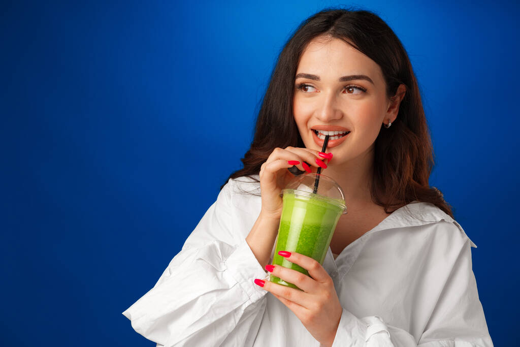 Młoda piękna kobieta w białej koszuli pije zielony smoothie na niebieskim tle - Zdjęcie, obraz