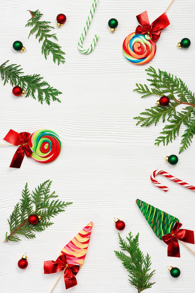 Dulces de Navidad surtidos vista superior, ramas de thuja verde, pequeños juguetes de bolas rojas y verdes sobre fondo de madera blanca, puesta plana. Dulces brillantes de Año Nuevo y piruletas. - Foto, imagen