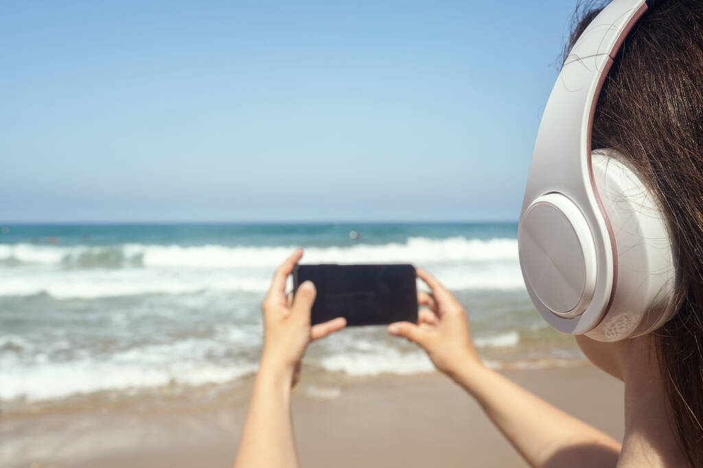 Samotna kobieta na plaży w słuchawkach słucha muzyki, fotografuje morze przez telefon. Kobiecy relaks na wakacjach. Widok z tyłu - Zdjęcie, obraz